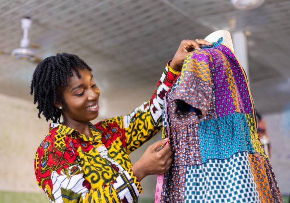 Smiling Ghanaian Dressmaker with locs hair measures a part of her colourful African pattern dress in her studio